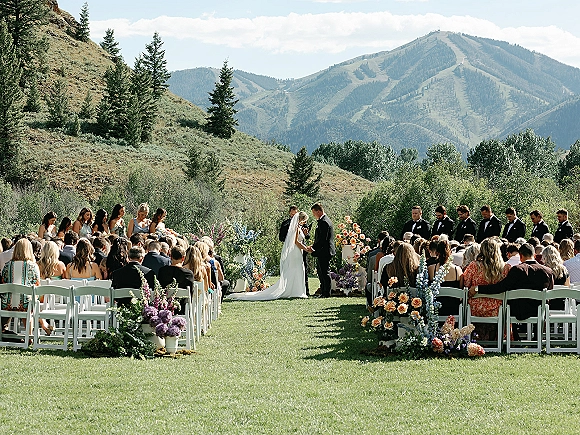 Wedding ceremony with bride and groom at the floral arch, guests in white chairs on a lawn with mountains and evergreens behind