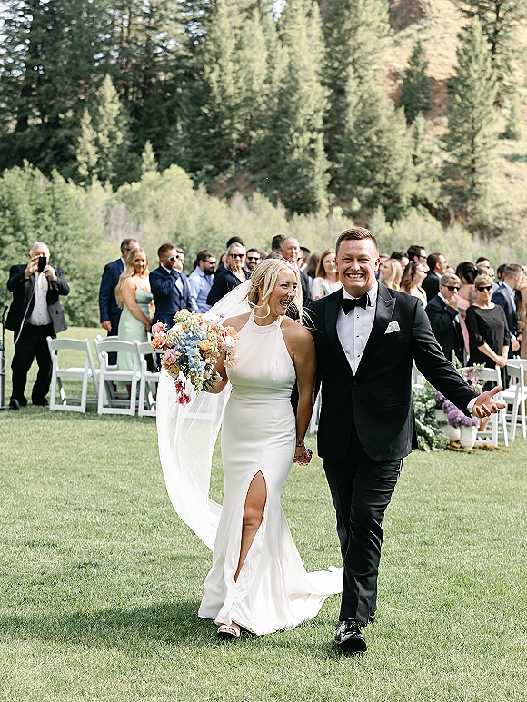 Wedding recessional as bride and groom walk the aisle holding hands, bride laughing with bouquet and long veil on an outdoor lawn with guests