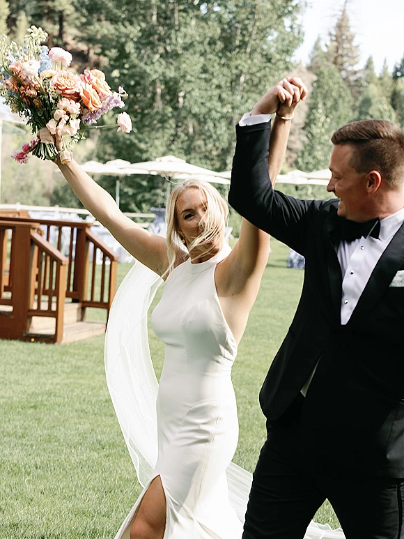 Newlywed celebration as bride and groom cheering hand in hand, bouquet raised, veil blowing on a sunny lawn by a wooden deck
