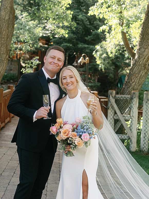 Couple portrait of bride and groom toasting with champagne, bride holding a colorful bouquet on a garden stone walkway with trees behind