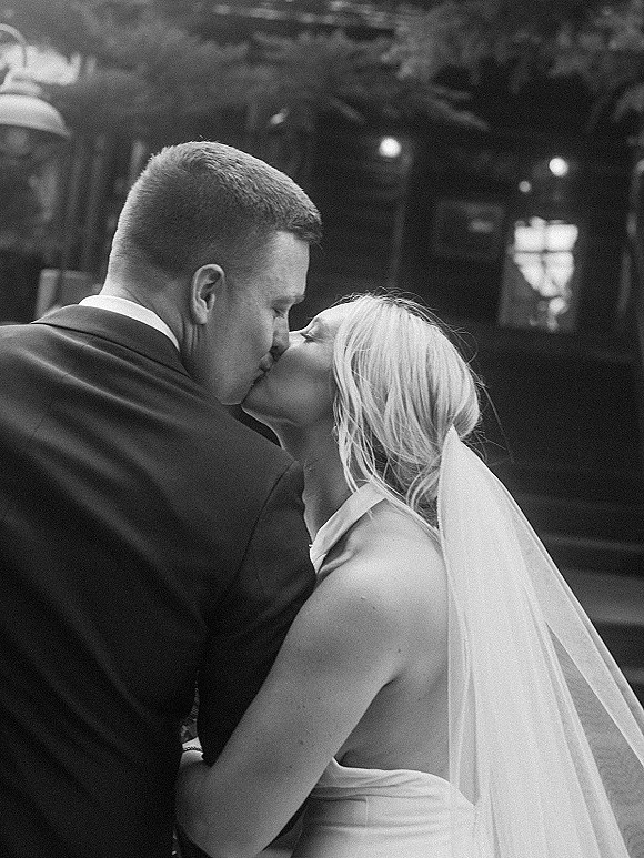 Wedding kiss as bride in strapless wedding dress and veil embraces groom with boutonniere on an outdoor patio under string lights