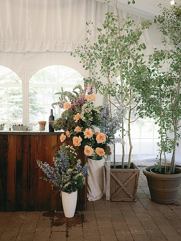 Wedding bar decor with peach and blue wedding flowers in a tall white vase on a rustic wood bar under a draped white tent with string lights