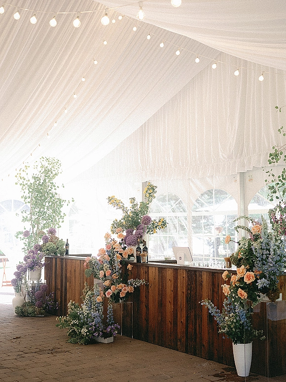 Wedding bar setup with rustic wedding bar facade, wine bottles and glassware beneath draped tent ceiling with string lights and florals