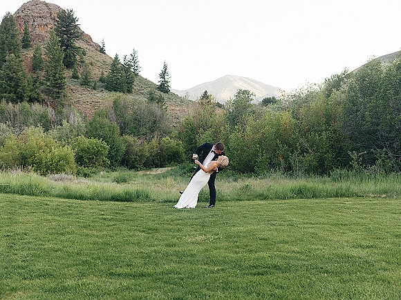 Wedding kiss portrait of groom dipping bride in a tuxedo and fitted dress, holding a champagne flute in a mountain meadow with pines