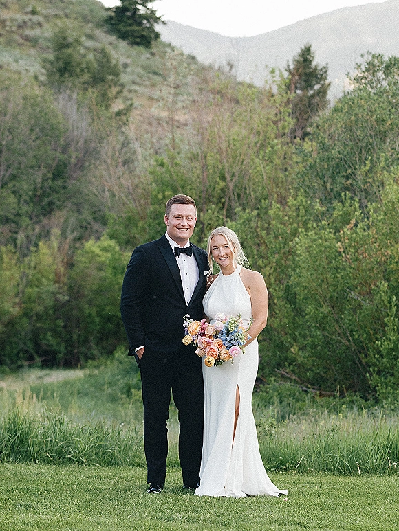 Couple portrait of bride and groom in a mountain wedding portrait, she holds a bridal bouquet in meadow grass with green hills behind