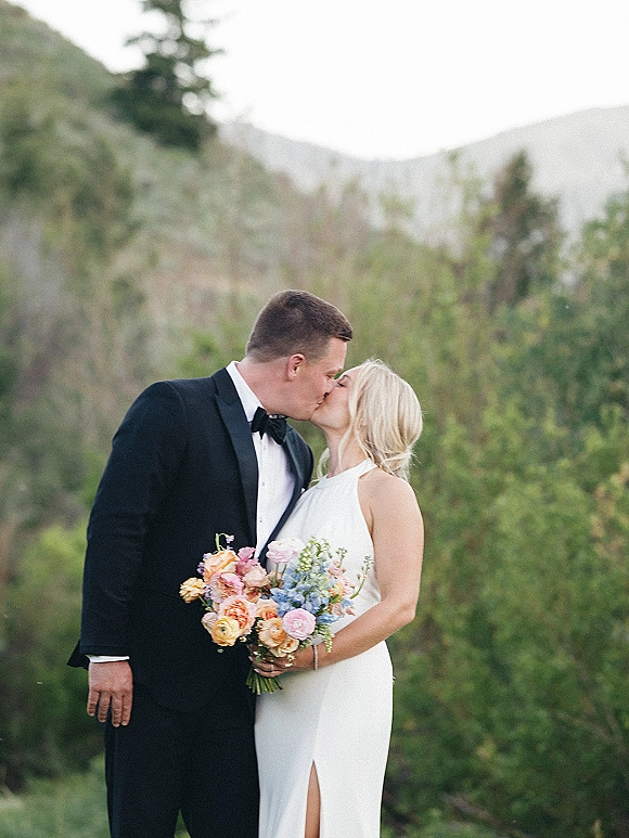 Wedding kiss portrait of bride and groom kissing, her colorful bouquet in hand, with mountains and greenery behind them in natural light