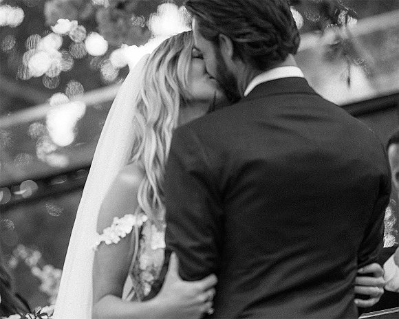 Wedding kiss as bride in a veil holds a bouquet while the groom in a dark suit embraces her under string lights with guests behind
