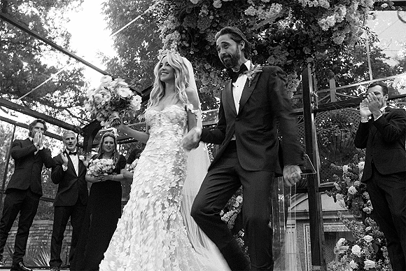 Wedding recessional as bride and groom holding hands walk back the aisle under a floral arch, guests applauding in an outdoor garden setting