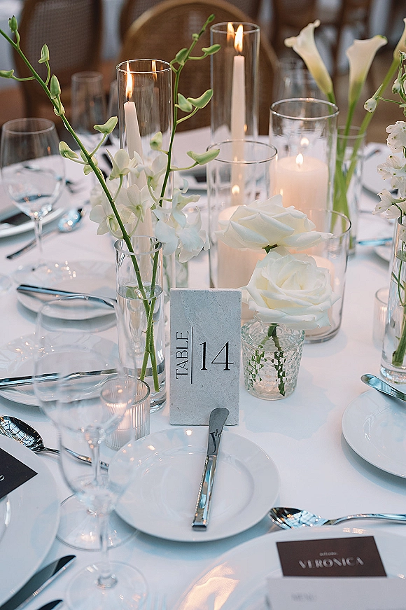 Reception tablescape with wedding table setting of white roses in bud vases, taper and pillar candles in glass hurricanes, place cards and table number.