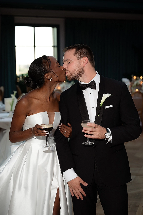 Wedding kiss portrait of bride and groom kissing, holding cocktail glasses in black tie attire by candlelit reception tables and window light
