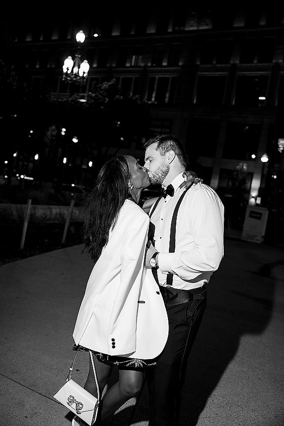 Wedding kiss portrait of a couple embracing on a city street at night under streetlights, bride in white blazer with a mini handbag