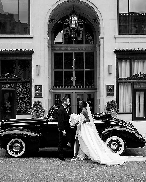 Couple portrait in a black and white wedding portrait, bride in veil holding bouquet facing groom in tuxedo beside a vintage convertible at a hotel entrance