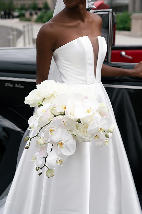 Bridal portrait of a bride in a strapless wedding dress with veil holding a white orchid bouquet beside a vintage car with open door