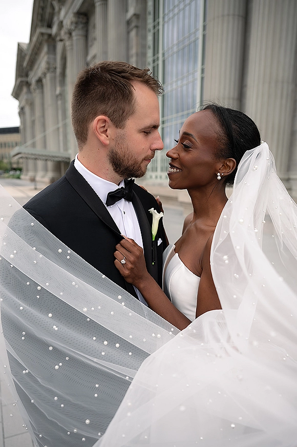 Couple portrait of bride and groom close up, forehead to forehead under a pearl wedding veil beside stone columns on a city sidewalk