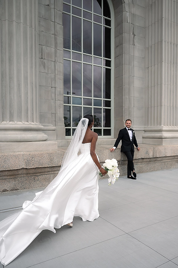 Wedding first look as bride walks to groom in a black tuxedo, her long veil and bouquet trailing by stone columns and arched window