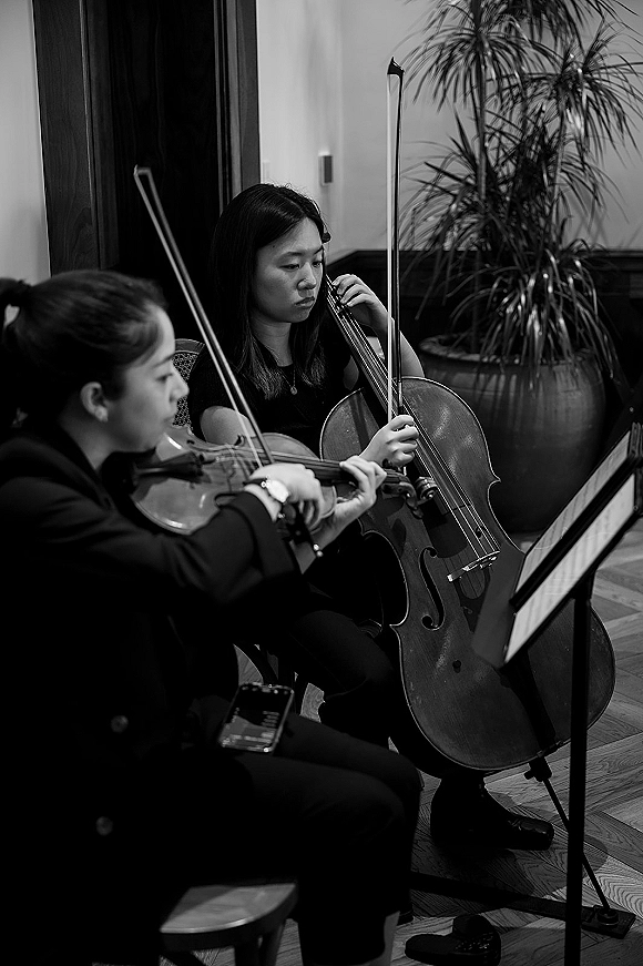 Wedding musicians, a wedding string duo playing violin and cello with sheet music on stands in an indoor venue with wood floors and paneling