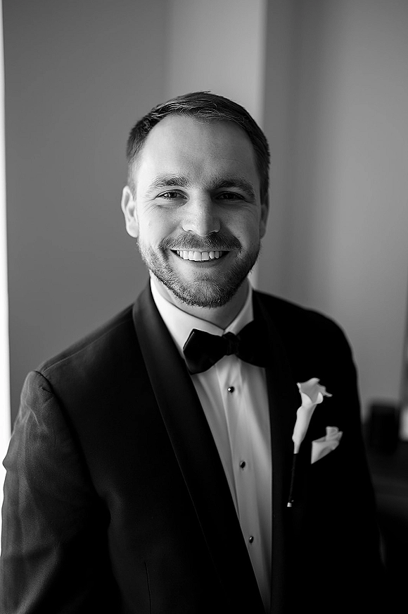 Groom portrait in a black tie tuxedo with bow tie and boutonniere, standing by a wall in soft window light indoors