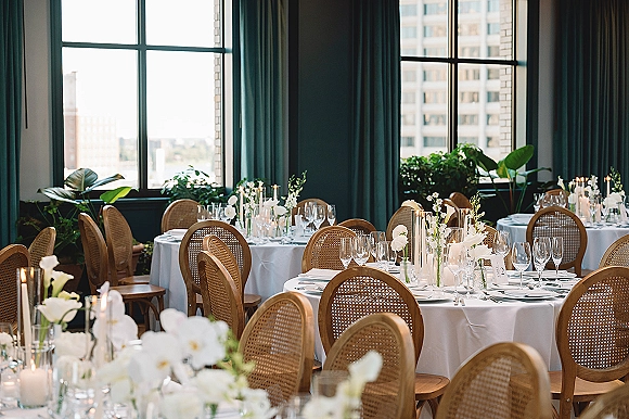 Reception tablescape with round wedding tables in white linens, cane back chairs, taper candles and white florals by large windows with city views