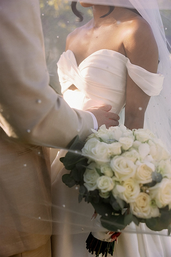 Couple portrait of bride and groom close up, her veil and white rose bouquet with greenery, his tan suit, in a sunlit garden