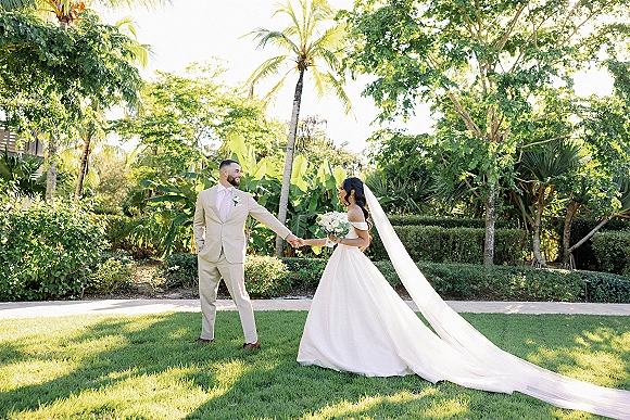 Couple portrait of bride and groom holding hands, her long veil and bouquet trailing as they stand on a sunlit tropical lawn with palm trees