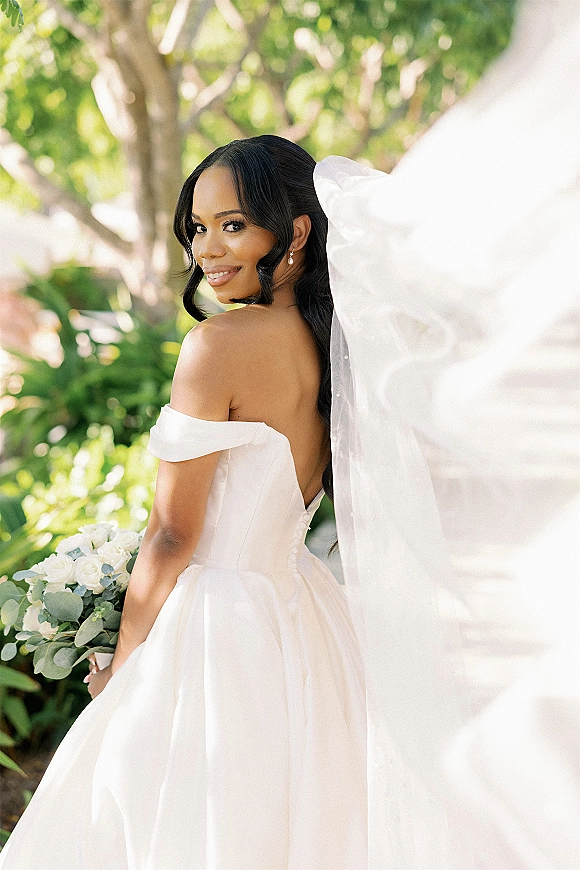 Bridal portrait of a bride looking back in an off the shoulder wedding dress with pearl earrings and a white rose bouquet in sunlit garden foliage