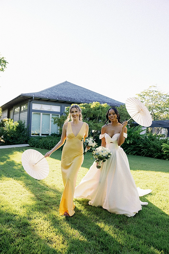 Bride and bridesmaid walking with bride under white paper parasols, holding white rose bouquets on a green lawn by a modern house exterior