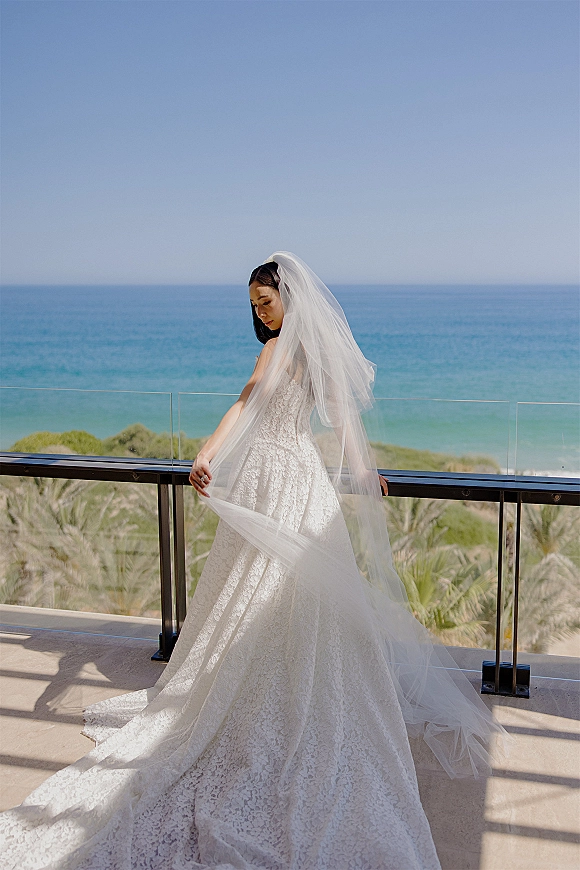 Bridal portrait of a bride in a lace wedding dress with cathedral veil, looking down on a balcony with ocean view and palm trees