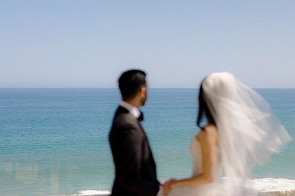 Couple portrait of bride and groom holding hands at a beach wedding portrait, seen from behind facing the ocean horizon with veil blowing
