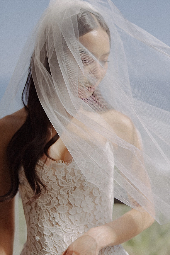 Bridal portrait of a bride under veil, looking down in a lace wedding dress against a blue sky in natural outdoor light