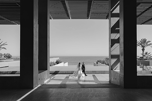 Couple portrait of bride and groom walking under concrete columns, her long train and veil trailing, with ocean horizon and palms beyond