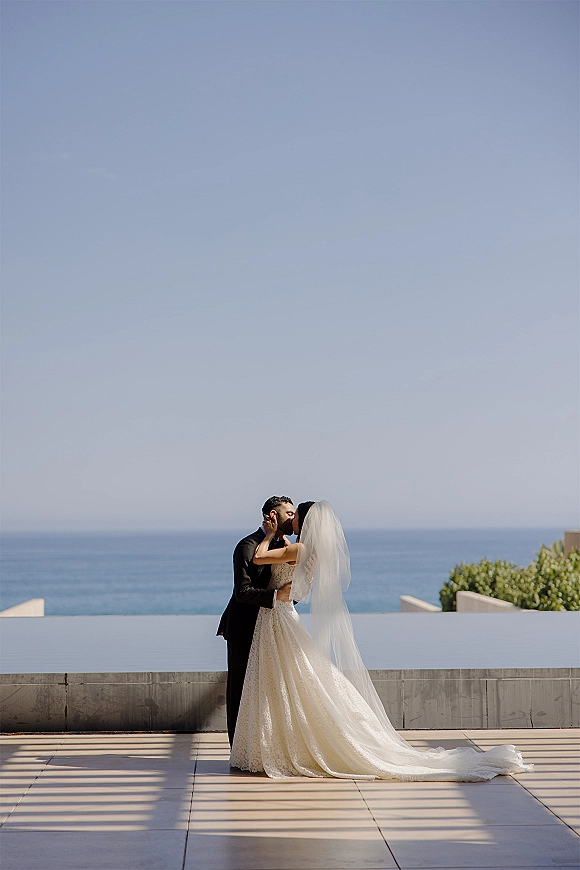 Wedding kiss portrait of bride and groom kissing, her long veil and lace dress trailing on a modern terrace with ocean horizon behind