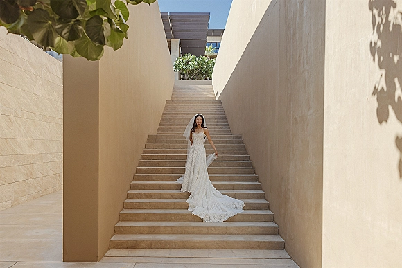 Bridal portrait of a bride in a strapless lace wedding dress with a cathedral veil, standing on a stone staircase by beige walls.