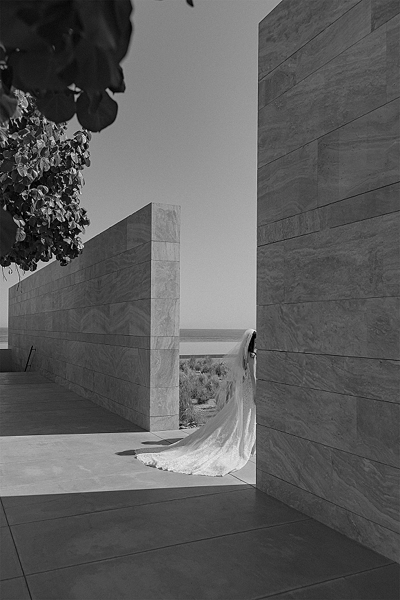 Bridal portrait of a bride in a long veil walking away, her wedding dress train flowing on a stone terrace with ocean horizon beyond