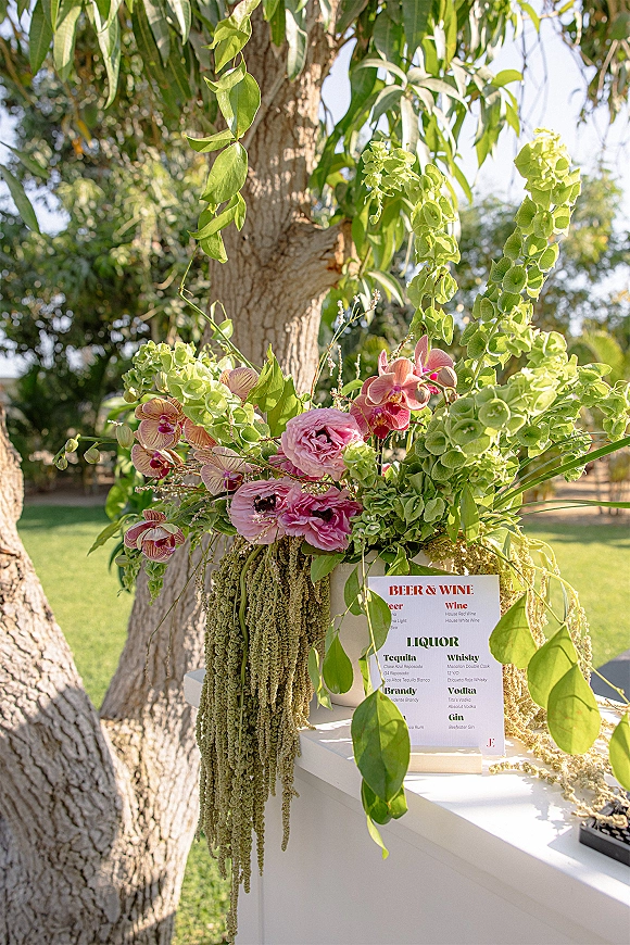 Bar menu sign for wedding bar menu framed by cascading orchids and pink roses with green hydrangea on a sunlit garden lawn near a tree trunk
