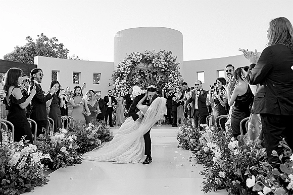 Wedding kiss as the groom dips the bride in her veil and gown beneath a floral arch, guests cheering in a white stucco courtyard.