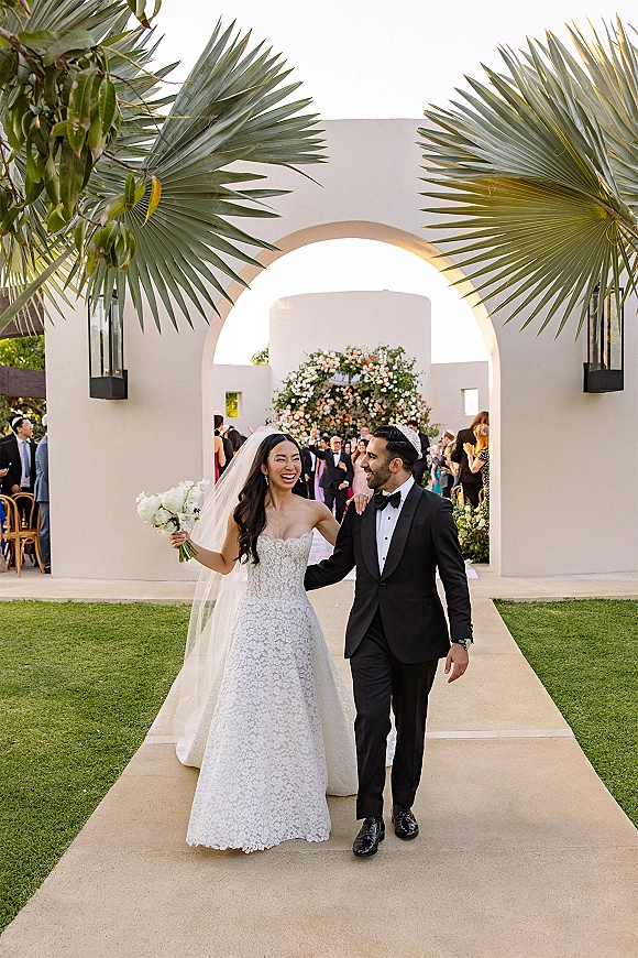 Wedding recessional as bride and groom walk the aisle laughing, her white rose bouquet and cathedral veil under a white stucco arch with palm fronds