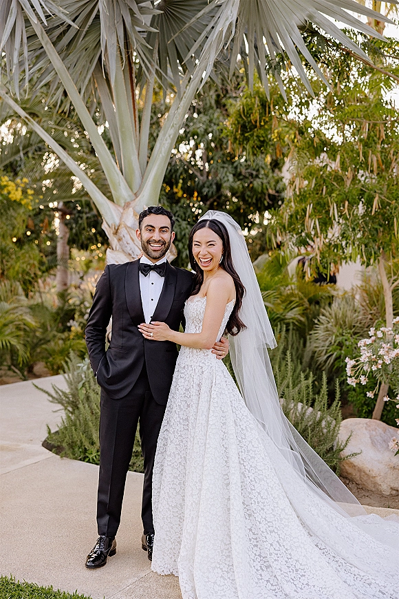 Couple portrait of bride and groom portrait laughing as she holds his lapel, with veil and tuxedo on a palm-lined garden pathway