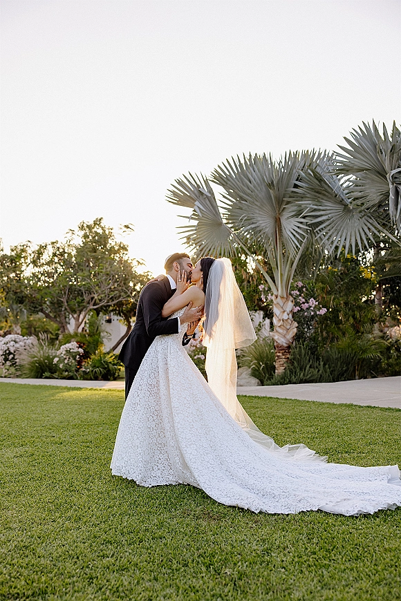 Wedding kiss portrait of bride and groom kissing, her lace dress and long veil trailing on a lawn with palm trees and flowering shrubs