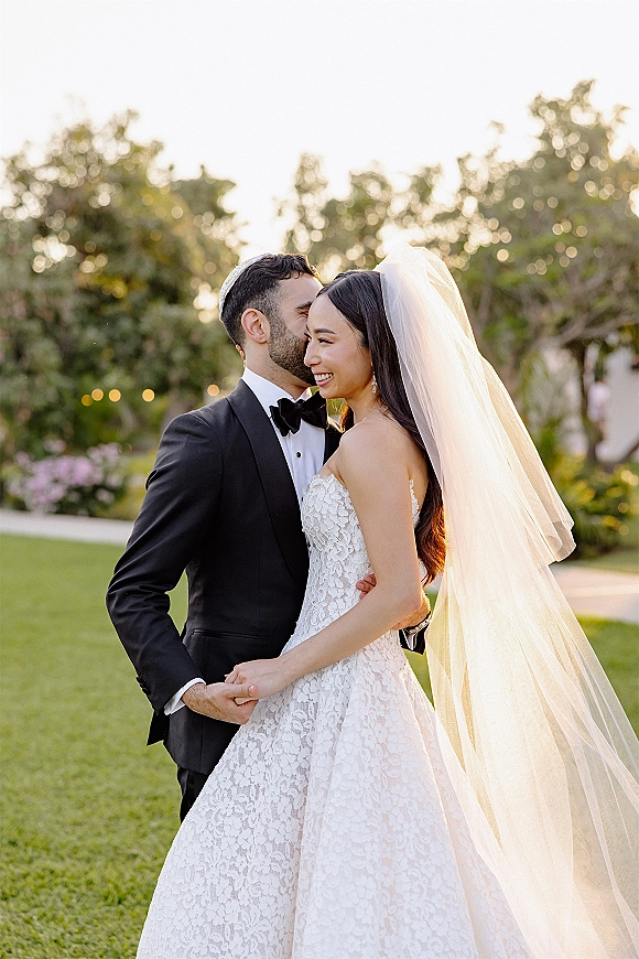 Couple portrait of bride and groom embrace, groom kissing her forehead as her wedding veil glows in soft garden sunlight