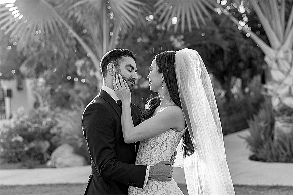 Couple portrait of bride touching groom’s face in a newlywed embrace, lace dress and veil flowing, with palm trees and greenery behind