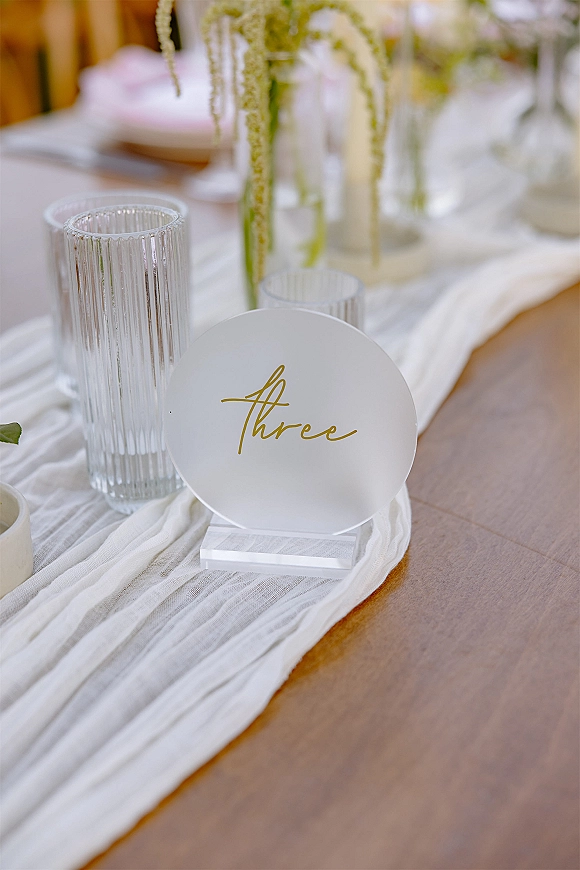 Wedding table number acrylic table number sign with gold calligraphy beside ribbed glass vase and votive on gauze runner, blurred reception decor