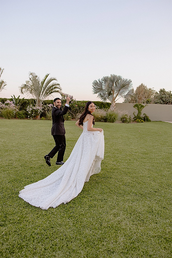 Couple portrait of bride and groom walking hand in hand, looking back, her lace dress train flowing on a palm-lined lawn at sunset