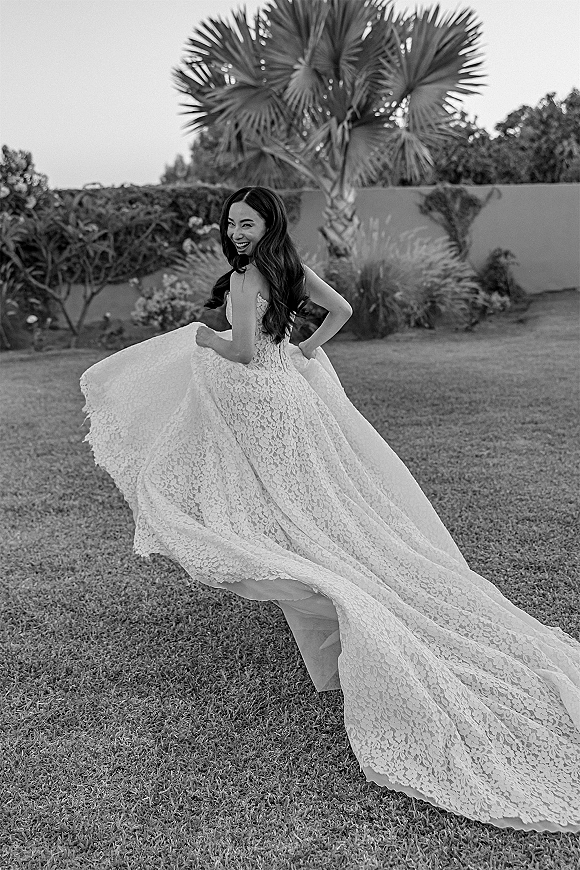 Bridal portrait of a bride twirling her strapless lace wedding dress, lifting the long train on grass with palm tree and shrubs behind