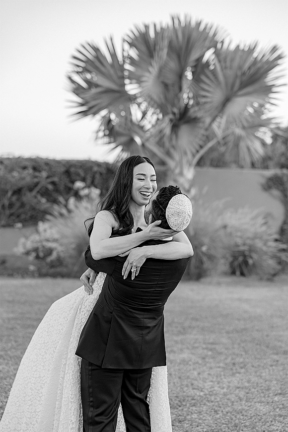 Couple portrait of groom lifting bride in a playful wedding portrait, her lace dress flowing as they laugh on a lawn by palm trees