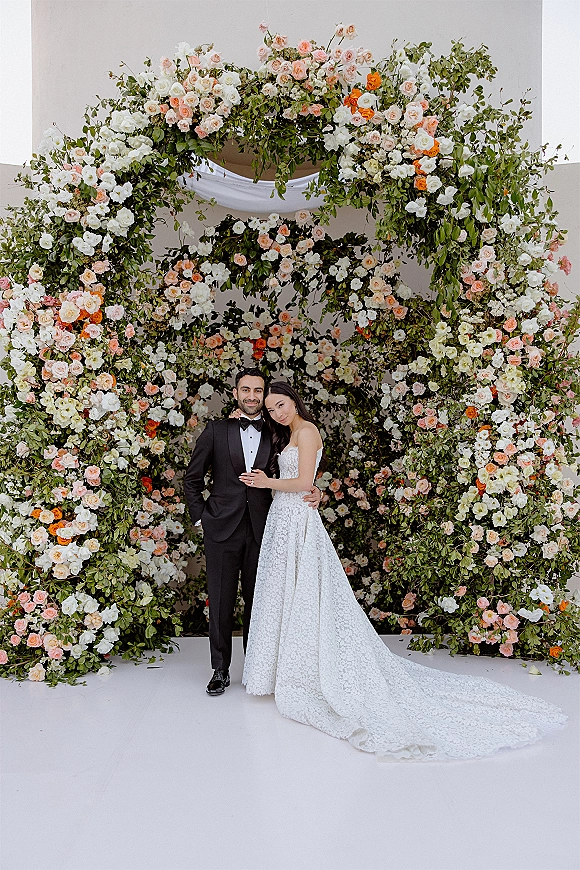 Couple portrait of bride in a strapless lace gown with long train and groom in tuxedo beneath a rose and greenery arch on white backdrop
