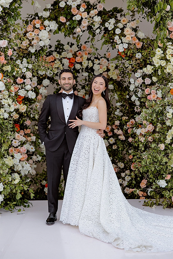 Couple portrait of bride in strapless lace gown laughing as groom in black tuxedo hugs her before a rose and greenery wall