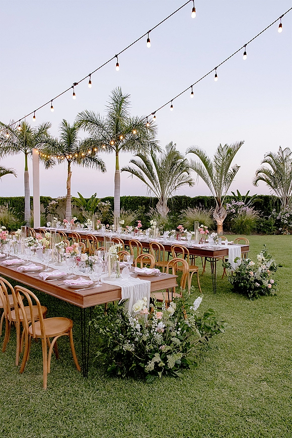 Outdoor reception tablescape with backyard wedding reception long wood tables, floral centerpieces and candlelight under string lights on a lawn with palm trees