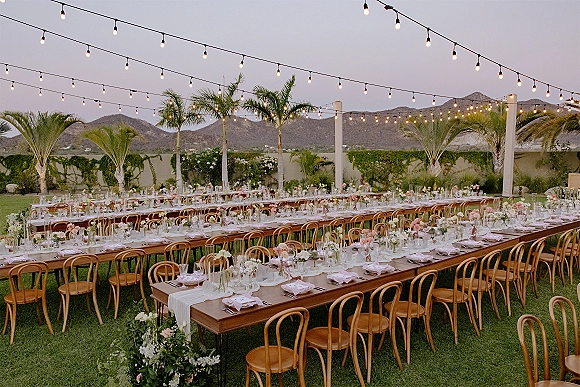 Reception tablescape on outdoor reception tables with blush napkins, taper candles and bud vase florals under string lights at dusk on a lawn with palms