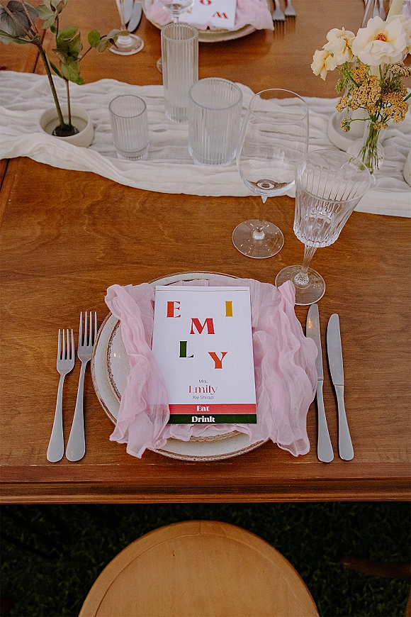 Wedding place setting with wedding menu card on stoneware plates, blush pink napkin, mix and match glassware, and rose bud vase on wooden table outdoors