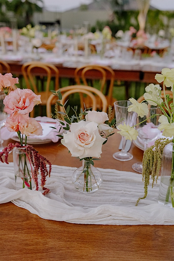 Reception tablescape with outdoor wedding reception table details, bud vases of roses, carnations, greenery on wood table with white runner on lawn